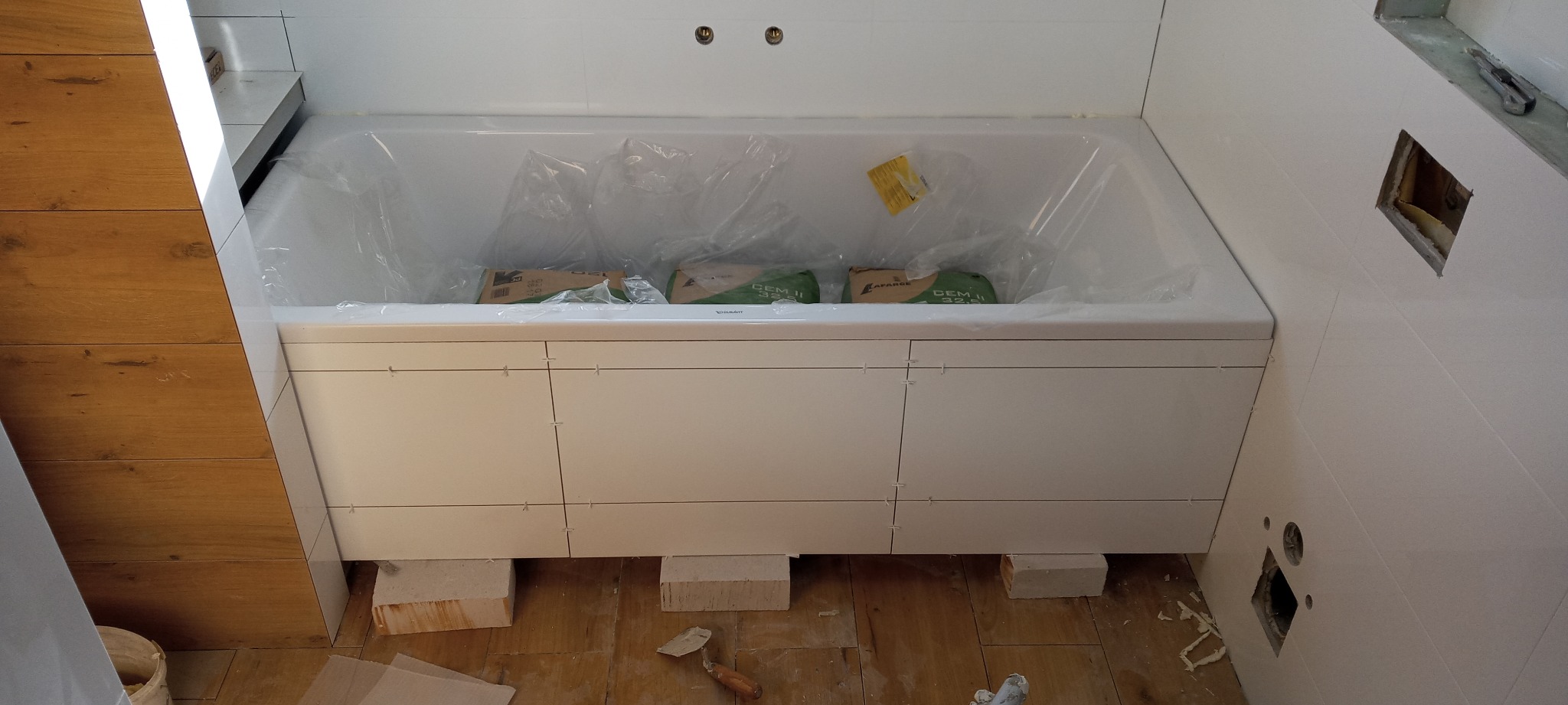 Biała wanna Duravit zabezpieczona folią, obudowana płytkami, podparta cegłami. Widoczne worki z zaprawą CEM II 32,5, szpachelka i płytki na podłodze. Ściany wyłożone białymi płytkami z wyciętym...