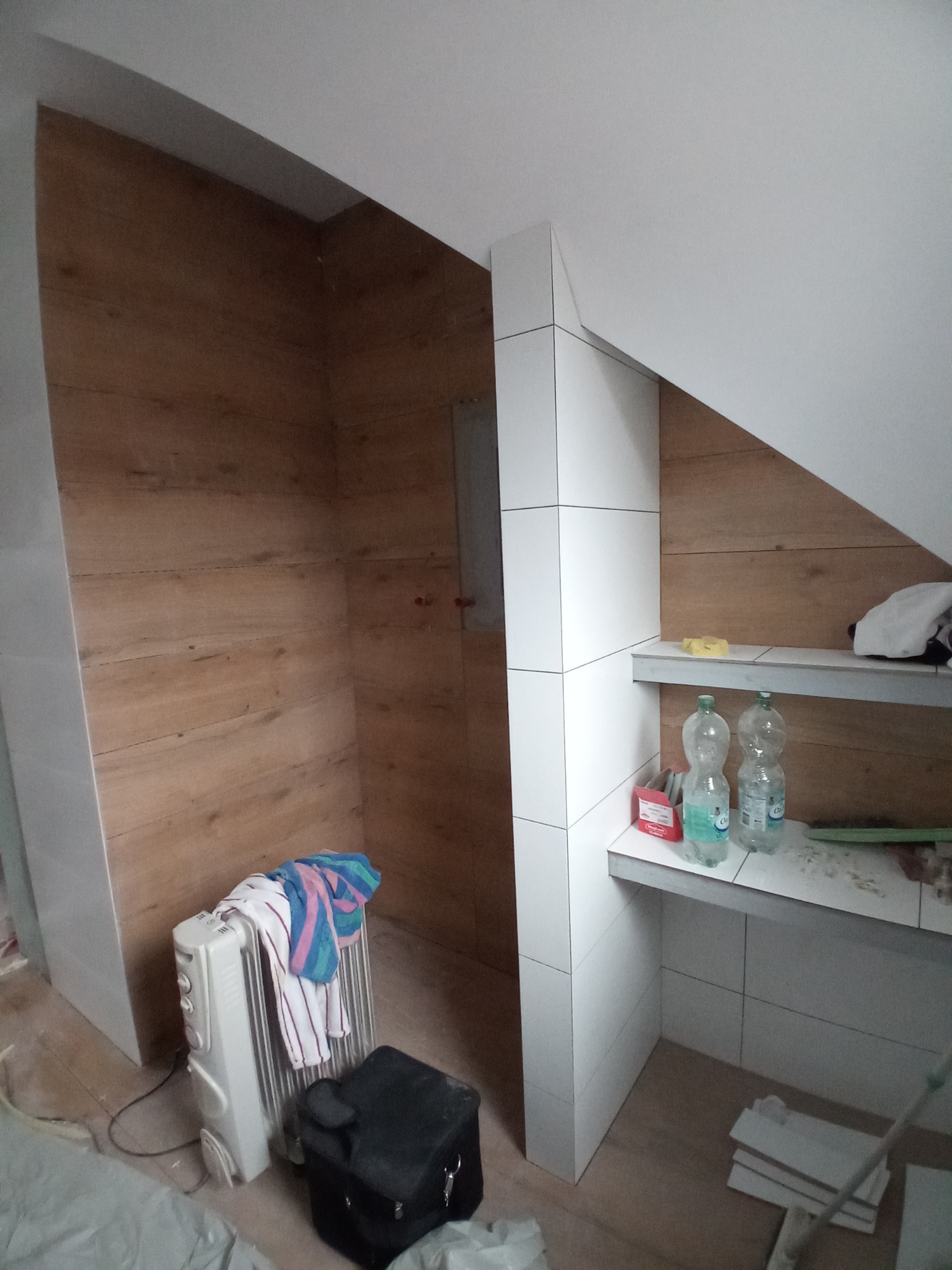 Adaptacja poddasza: ściana wykończona panelami imitującymi drewno, fragment zabudowy z białych płytek, grzejnik elektryczny z suszącymi się ręcznikami, butelki z wodą na półce.