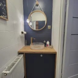 Nowoczesna łazienka z okrągłym lustrem zawieszonym na sznurku, szklaną umywalką na drewnianym blacie, ciemnogranatową ścianą i białymi płytkami z subtelnym wzorem.