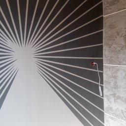 Paweł Barylka - Ściana pomalowana w geometryczny wzór promieni w kolorach szarym i białym, z widocznym fragmentem dekoracyjnej ściany obok i wystającym przewodem z żarówką.