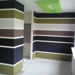 Paweł Barylka - Wnętrze pokoju z pomalowanymi ścianami w poziome pasy w odcieniach czerni, brązu, oliwki i bieli, z zielonym podświetlanym sufitem.