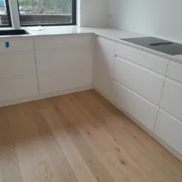 Minimalistyczna, biała zabudowa kuchenna z blatem z jasnego konglomeratu i płytą indukcyjną, widok na drewnianą podłogę i okno z zielenią w tle.