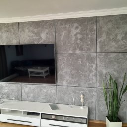 Paweł Barylka - Ściana w Kozienicach wykończona dekoracyjnym tynkiem imitującym marmur, podzielona na panele z ciemnymi fugami, z wiszącym telewizorem i białą szafką RTV z dekoracjami.