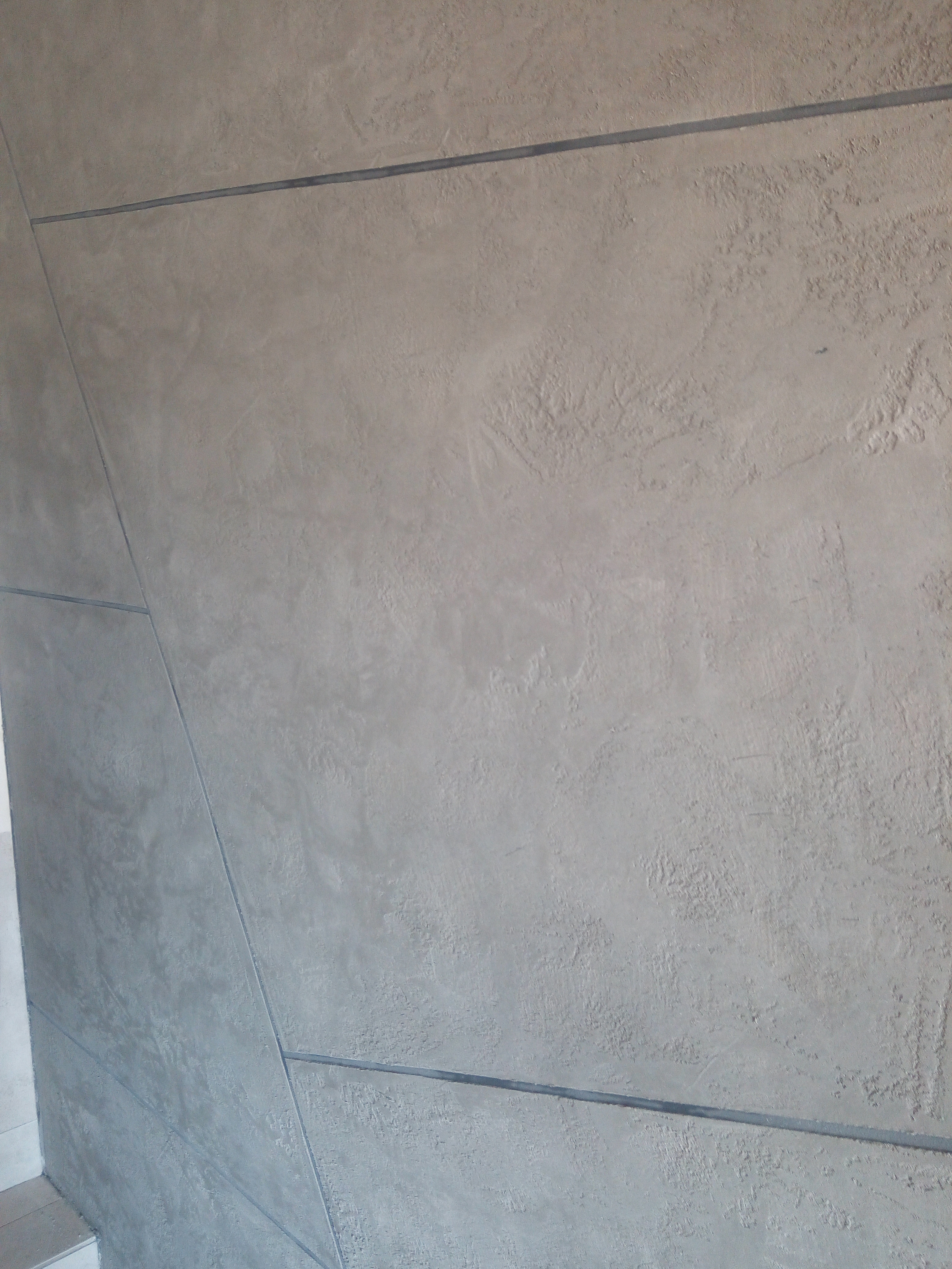 Dekoracyjna ściana w odcieniach szarości z poziomymi i ukośnymi liniami oddzielającymi fragmenty o różnej fakturze.