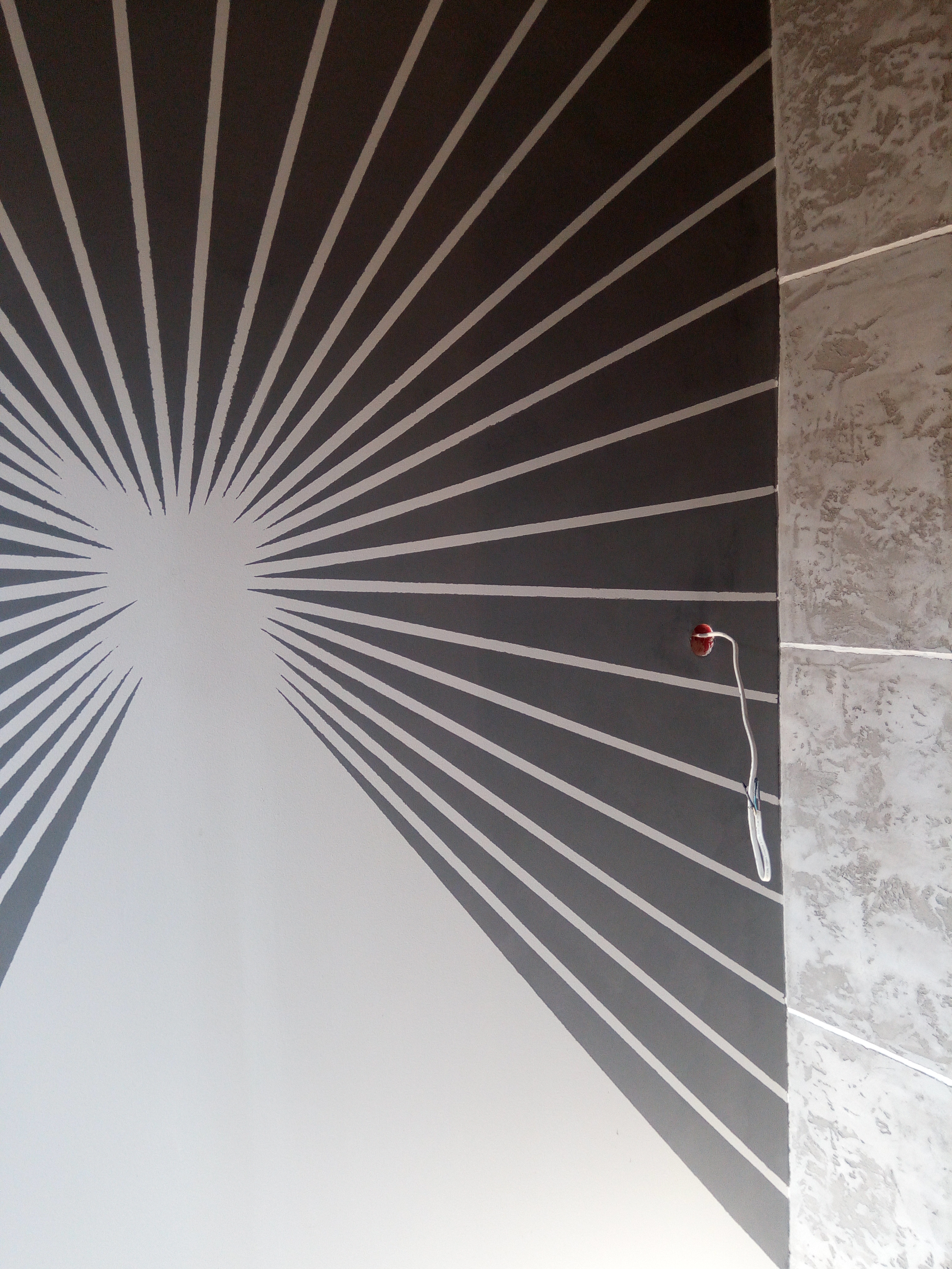 Ściana pomalowana w geometryczny wzór promieni w kolorach szarym i białym, z widocznym fragmentem dekoracyjnej ściany obok i wystającym przewodem z żarówką.
