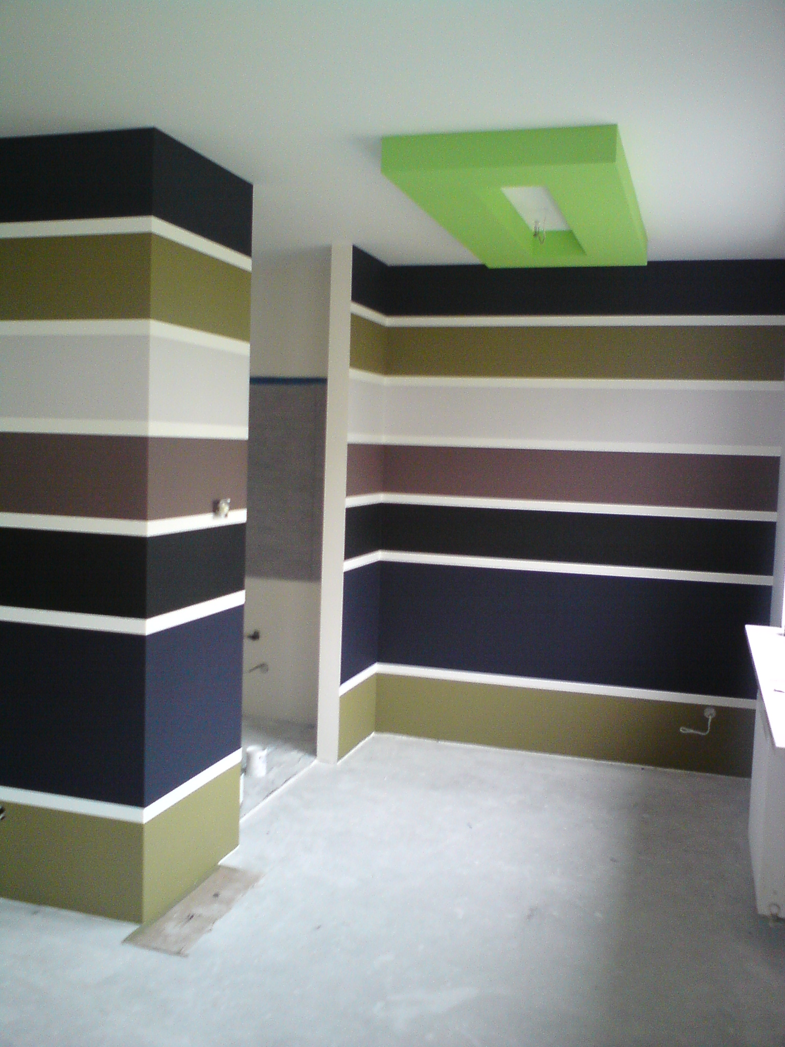 Wnętrze pokoju z pomalowanymi ścianami w poziome pasy w odcieniach czerni, brązu, oliwki i bieli, z zielonym podświetlanym sufitem.