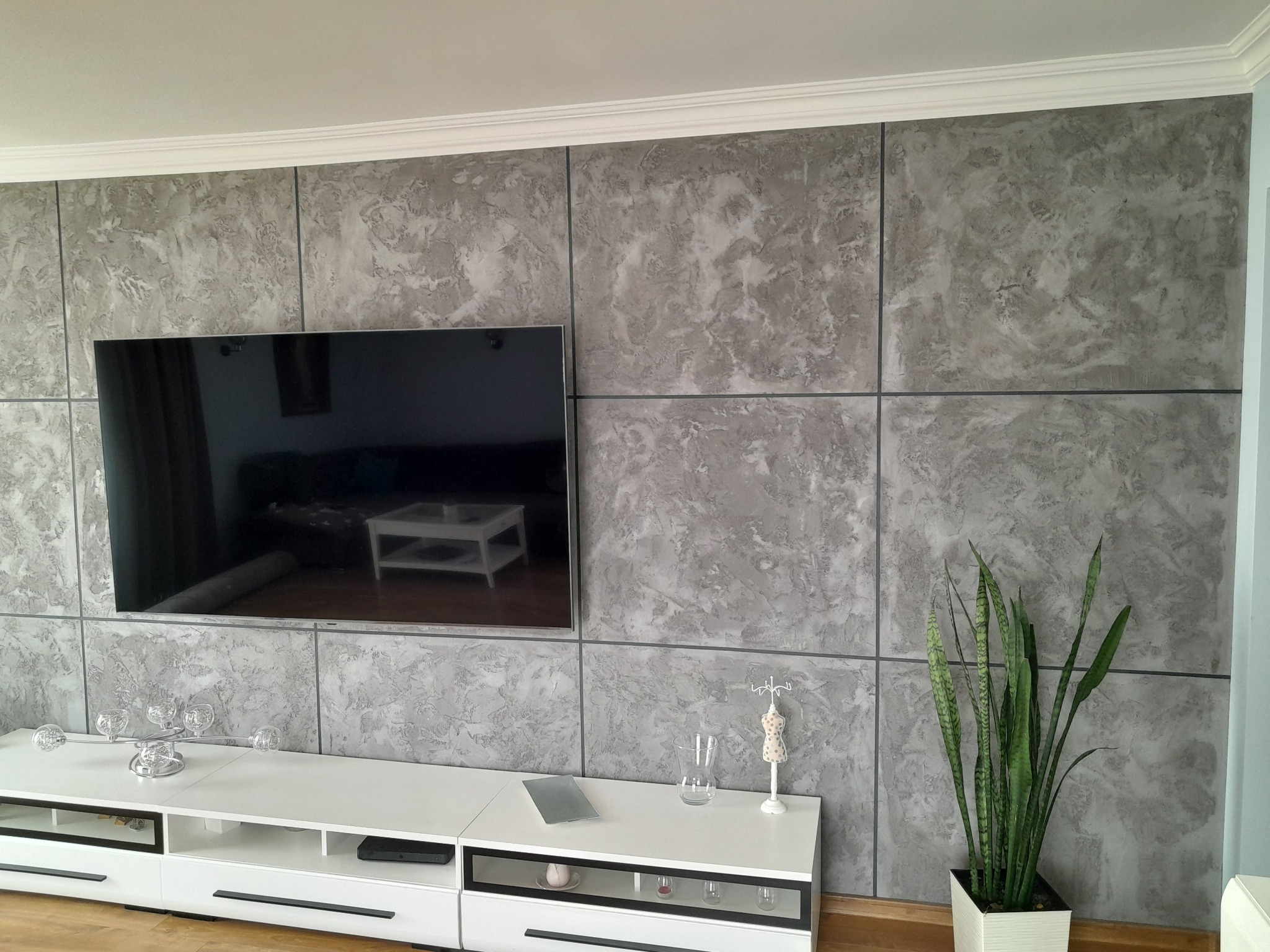 Ściana w Kozienicach wykończona dekoracyjnym tynkiem imitującym marmur, podzielona na panele z ciemnymi fugami, z wiszącym telewizorem i białą szafką RTV z dekoracjami.