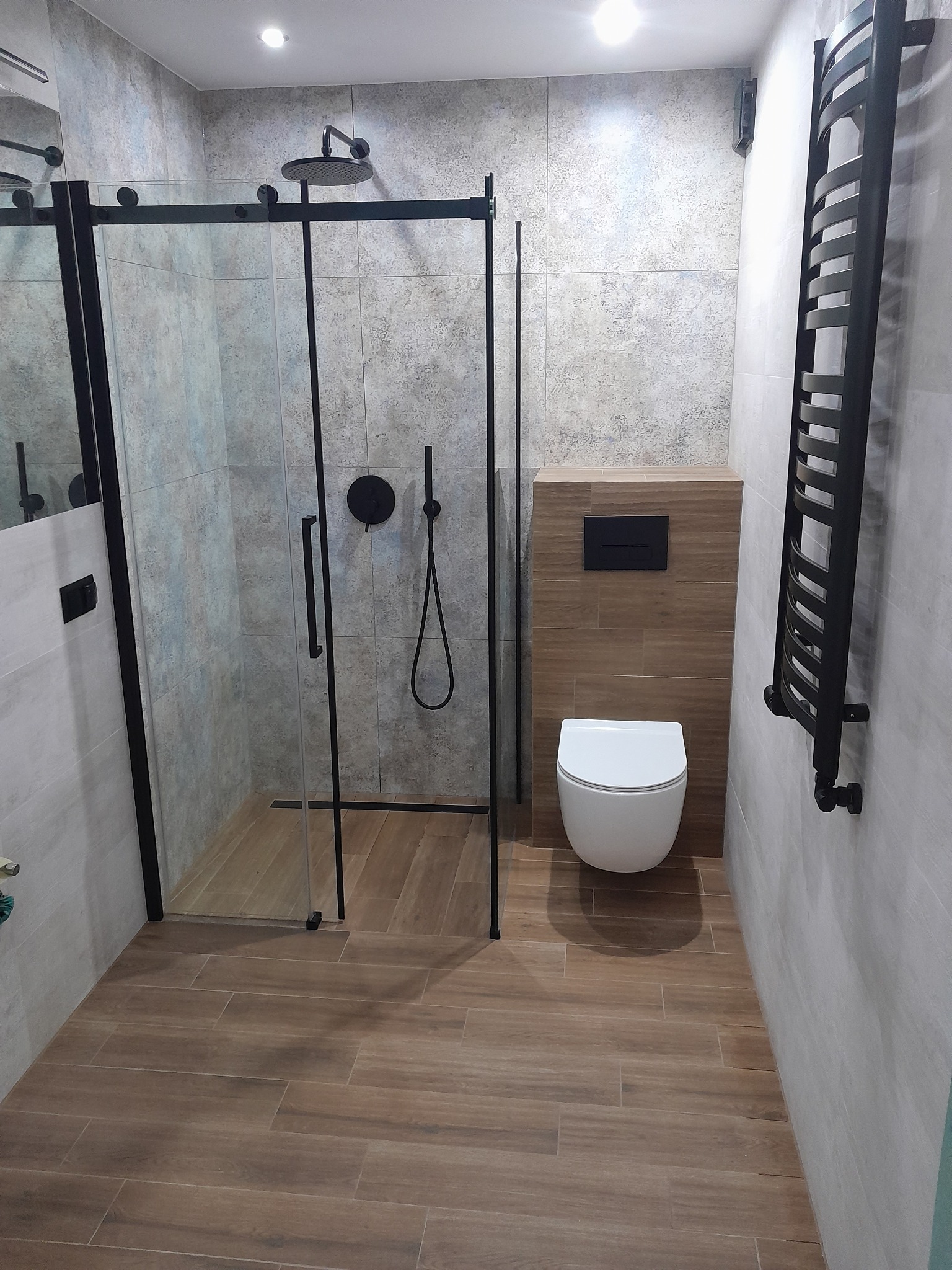 Nowoczesna łazienka z kabiną prysznicową typu walk-in z czarnymi profilami, wiszącą toaletą i grzejnikiem drabinkowym w kolorze czarnym, wykończona płytkami imitującymi drewno na podłodze i szarymi...