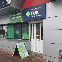 Fasada budynku z szyldem 'oferte dostarcza CUK UBEZPIECZENIA' nad wejściem, obok szyld 'ZOOLOGICZNO - WĘDKARSKI', okna z reklamami OC/AC i stojak reklamowy przed wejściem 'OBNIŻYMY CENĘ TWOJEGO...