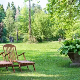 Dwa drewniane leżaki ogrodowe z podnóżkami na trawniku, w tle zieleń drzew i krzewów, duża betonowa donica z roślinami po prawej.