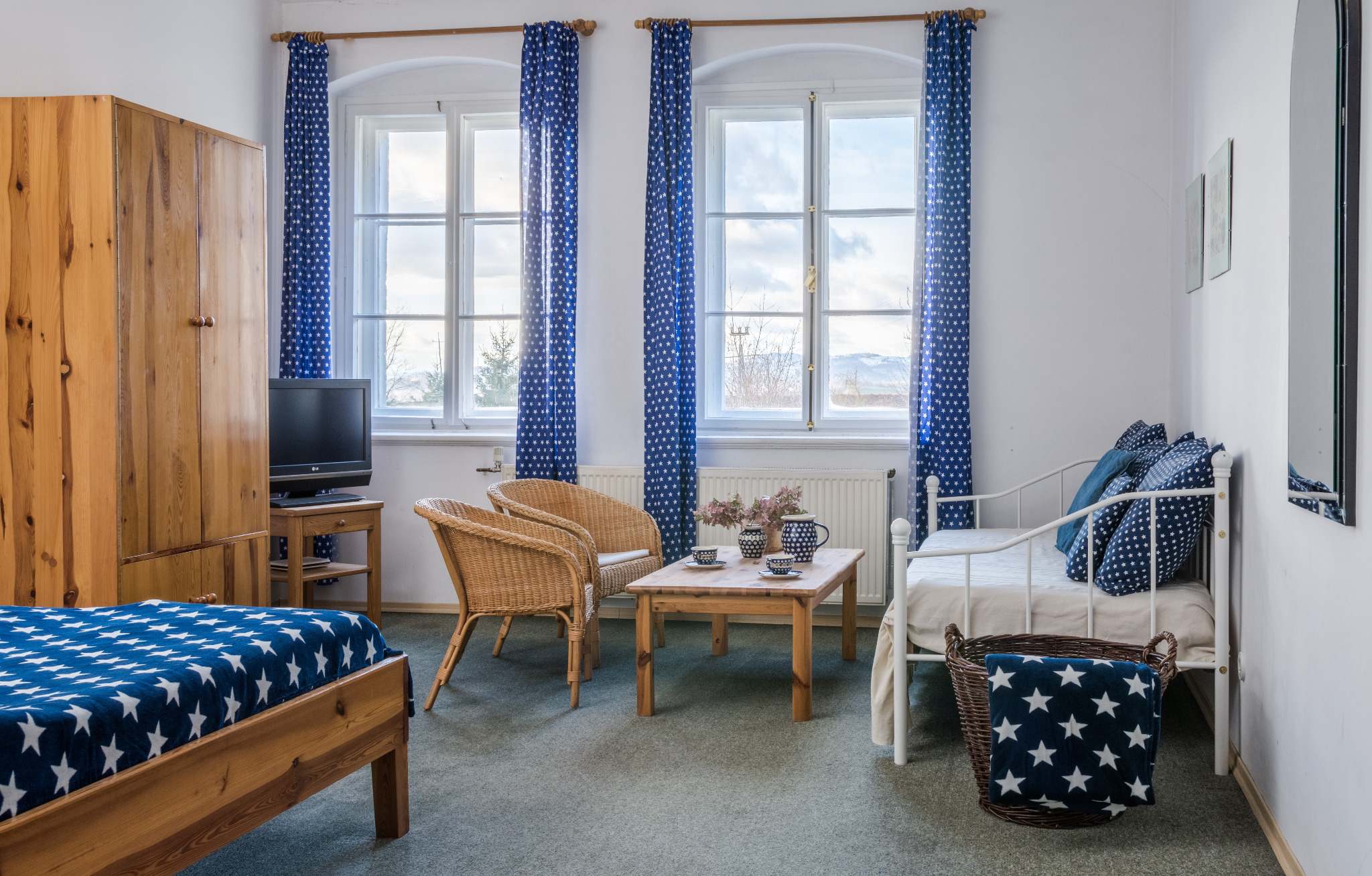 Jasny pokój gościnny z drewnianymi meblami, oknami z niebieskimi zasłonami w białe gwiazdki i łóżkami z pościelą w tym samym wzorze, na stoliku serwis do herbaty.