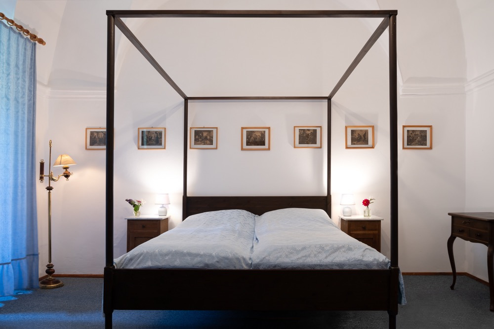 Pokój hotelowy z łóżkiem z baldachimem, lampką nocną i kwiatami, udekorowany grafikami na ścianie.