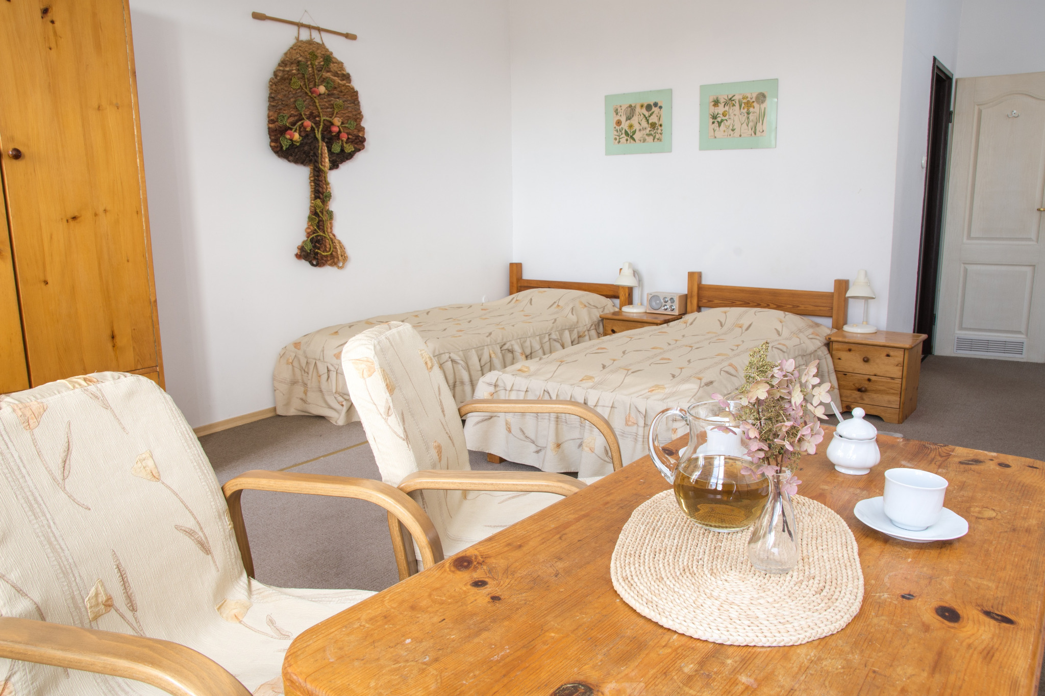 Pokój gościnny z dwoma łóżkami pojedynczymi, drewnianymi szafkami nocnymi i dekoracjami ściennymi, na pierwszym planie drewniany stół z dzbankiem herbaty i filiżanką.