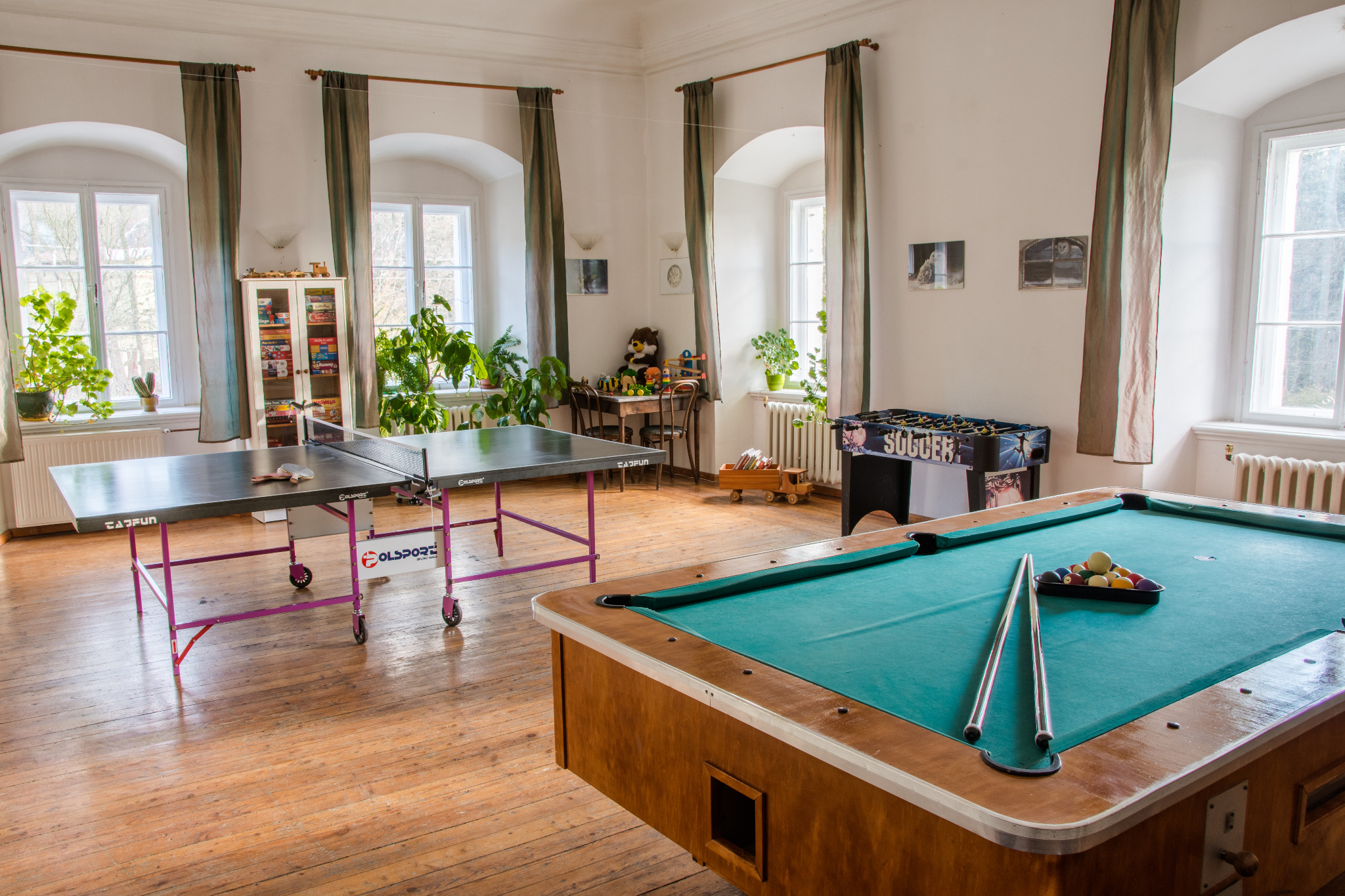 Sala gier z dużymi oknami, stołem bilardowym z kijami i trójkątem z bilami, stołem do ping-ponga z rakietkami, piłkarzykami i regałem z grami planszowymi.