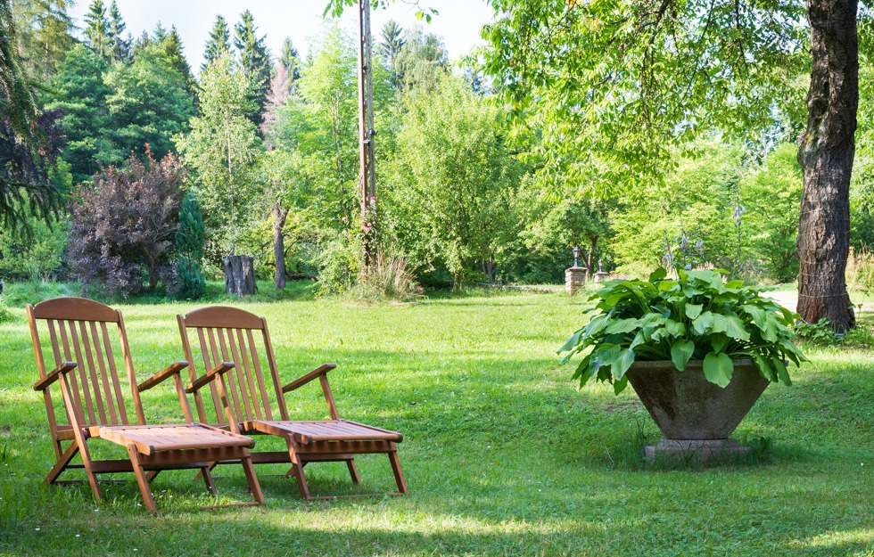 Dwa drewniane leżaki ogrodowe z podnóżkami na trawniku, w tle zieleń drzew i krzewów, duża betonowa donica z roślinami po prawej.