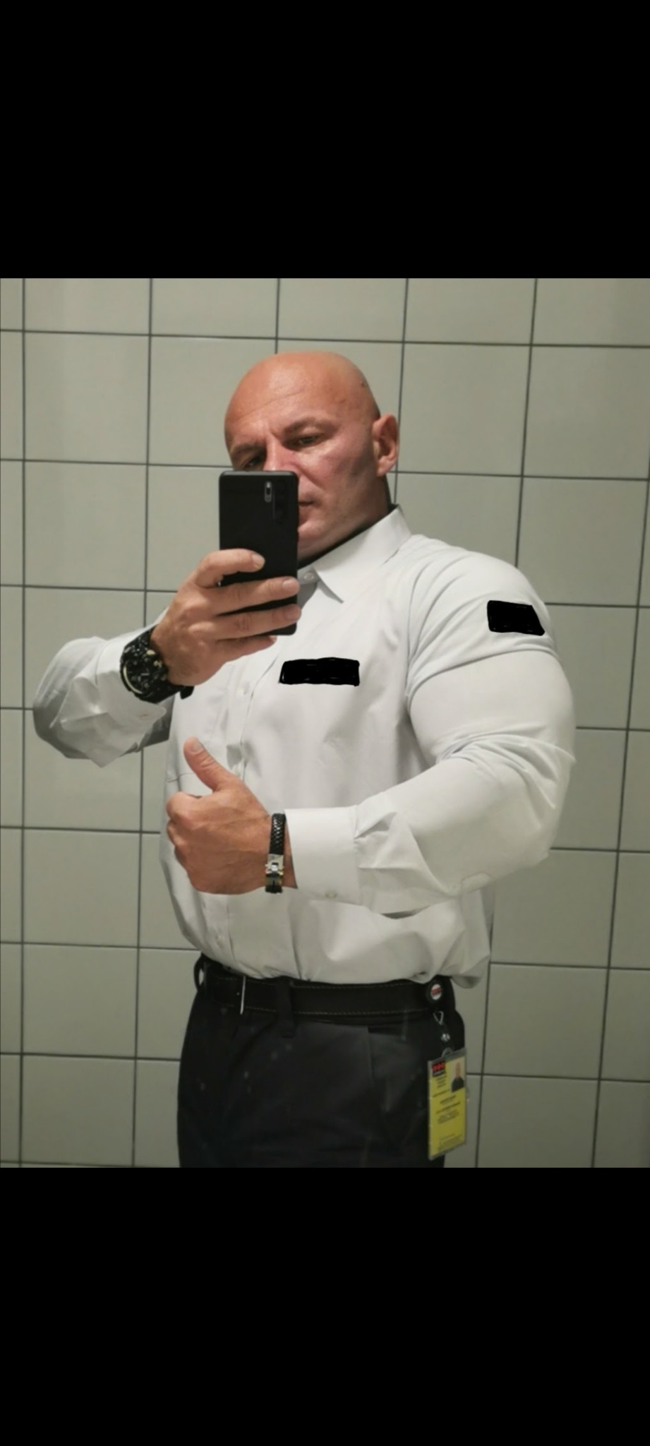 Mężczyzna w białej koszuli i ciemnych spodniach robi sobie selfie w lustrze na tle kafelków, pokazując kciuk w górę.