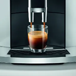 Automatyczny ekspres do kawy w trakcie przygotowywania espresso, z widocznymi dwoma strumieniami kawy wpadającymi do szklanki, na tle czarnego panelu i srebrnej obudowy.