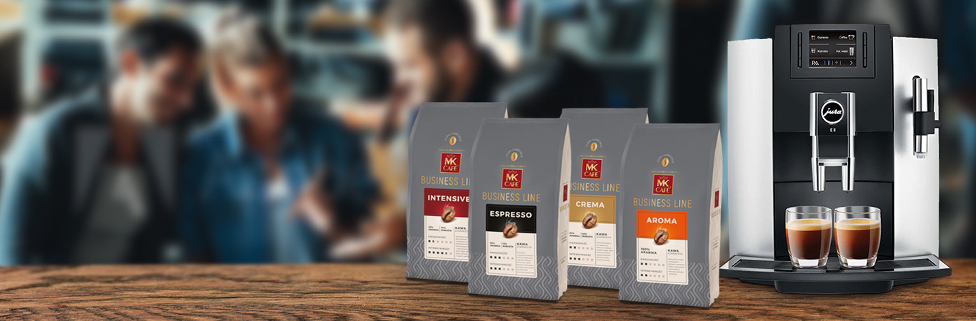 Automatyczny ekspres do kawy Jura E8 z dwoma szklankami espresso na tacy, obok cztery opakowania kawy MK Cafe Business Line: Intensive, Espresso, Crema i Aroma, w tle rozmyte postacie osób w biurze.