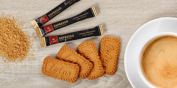 Kompozycja z filiżanką kawy, ciasteczkami Lotus, trzema saszetkami cukru trzcinowego MK Cafe Espresso Professional i kupką cukru trzcinowego na jasnym drewnianym tle.
