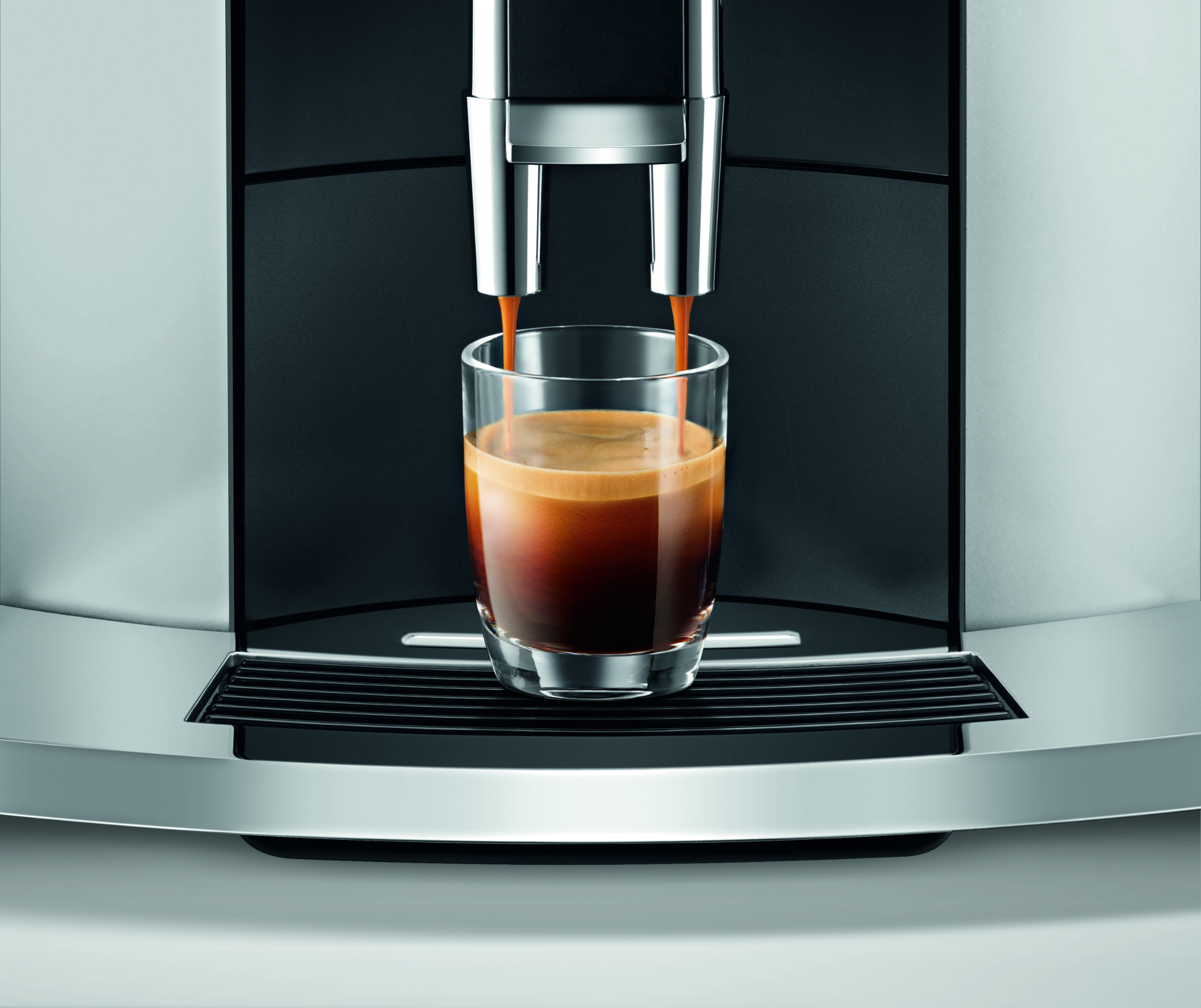 Automatyczny ekspres do kawy w trakcie przygotowywania espresso, z widocznymi dwoma strumieniami kawy wpadającymi do szklanki, na tle czarnego panelu i srebrnej obudowy.