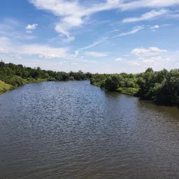 Szeroki kadr rzeki wijącej się między brzegami porośniętymi zielenią, pod błękitnym niebem z delikatnymi obłokami, widok z góry.