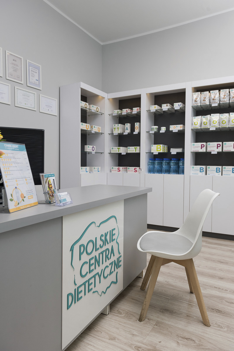 Wnętrze gabinetu dietetycznego z ladą recepcyjną, logo 'Polskie Centra Dietetyczne', regałami z produktami i cennikiem usług.