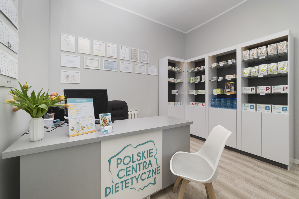 Jasne wnętrze gabinetu dietetycznego z biurkiem, kwiatami w wazonie, cennikiem usług i regałami z suplementami diety. Na ścianie wiszą certyfikaty.