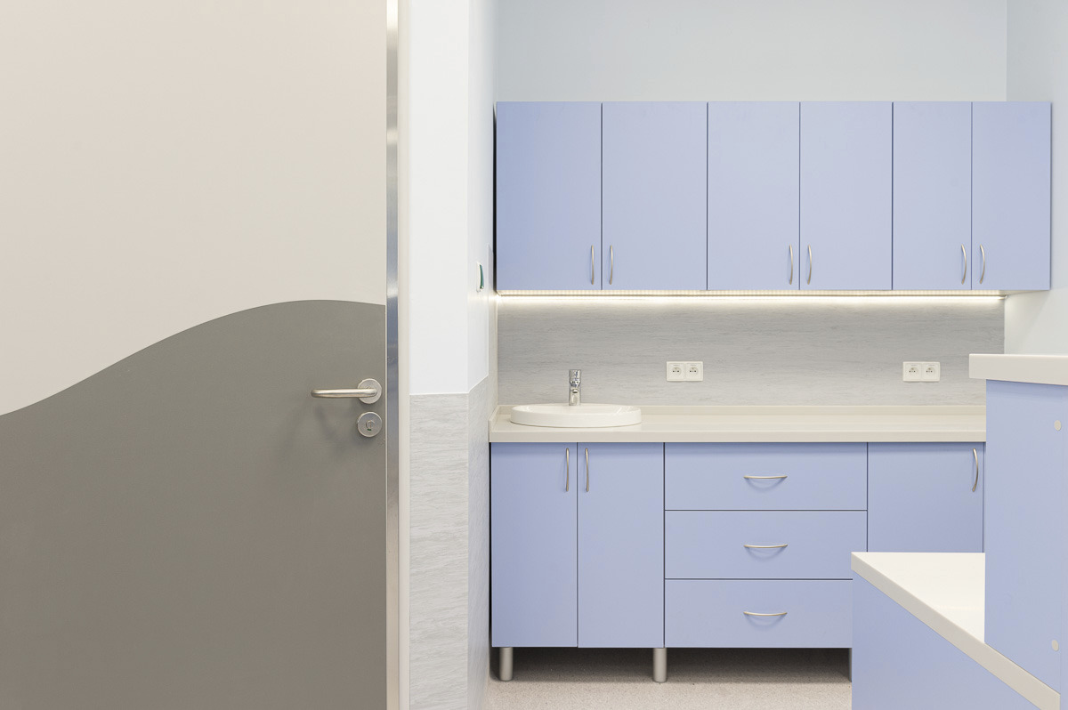 Jasne, nowoczesne wnętrze: błękitne szafki kuchenne z chromowanymi uchwytami, biały blat z umywalką, podświetlenie LED, szaro-białe marmurowe płytki na ścianie i dwukolorowe drzwi z srebrnym...