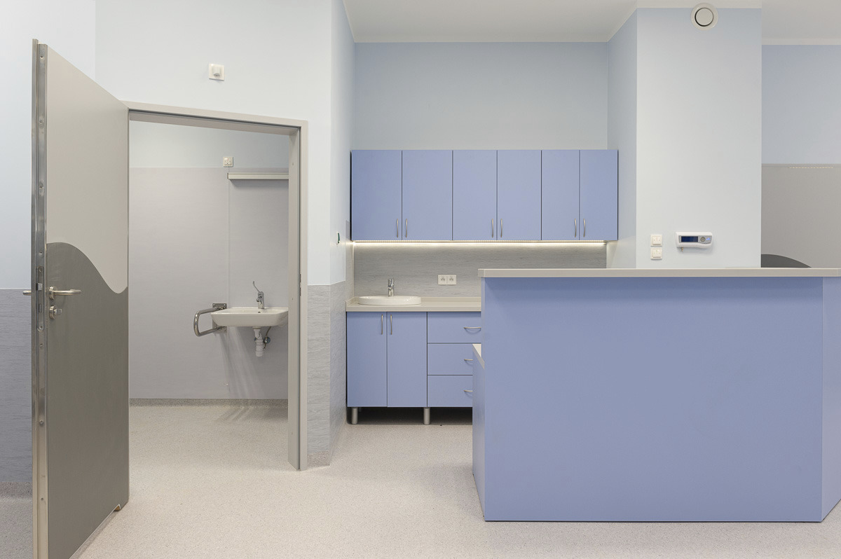 Jasne wnętrze gabinetu lekarskiego z niebieskimi szafkami, umywalką i ladą recepcyjną, widok na toaletę z umywalką przez otwarte drzwi.