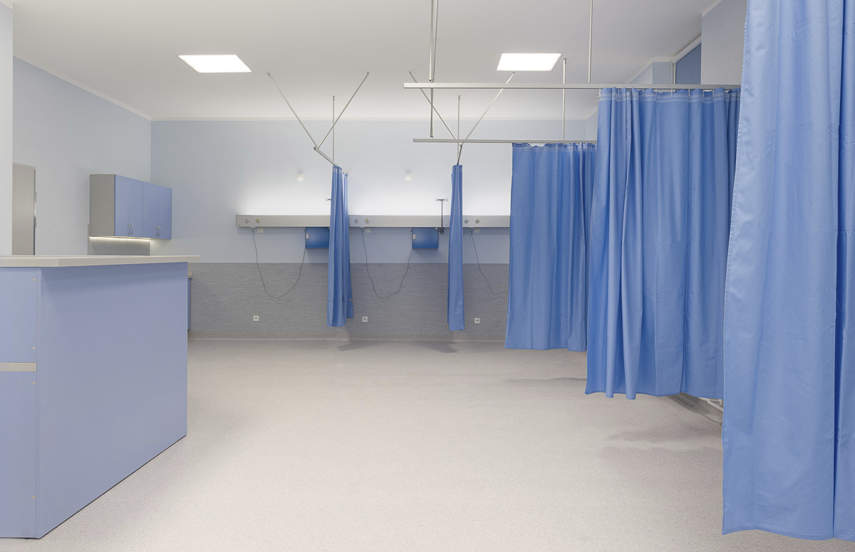 Pusta, jasna poczekalnia lub gabinet lekarski z niebieskimi zasłonami, biurkiem recepcyjnym i szafkami, oświetlona lampami sufitowymi.