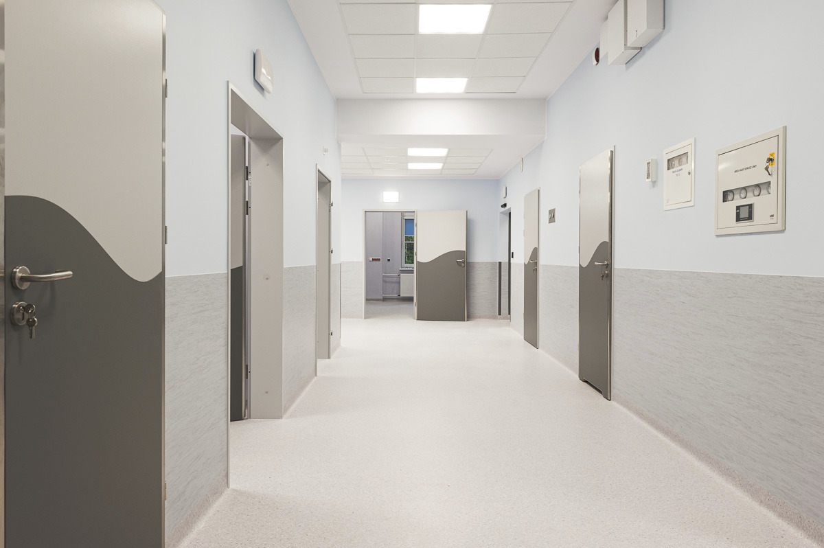 Długi, jasny korytarz w nowoczesnym budynku z rzędem szarych drzwi z dekoracyjnym wzorem fali, białe ściany z szarym panelowaniem, biała podłoga i sufit z kwadratowymi lampami.