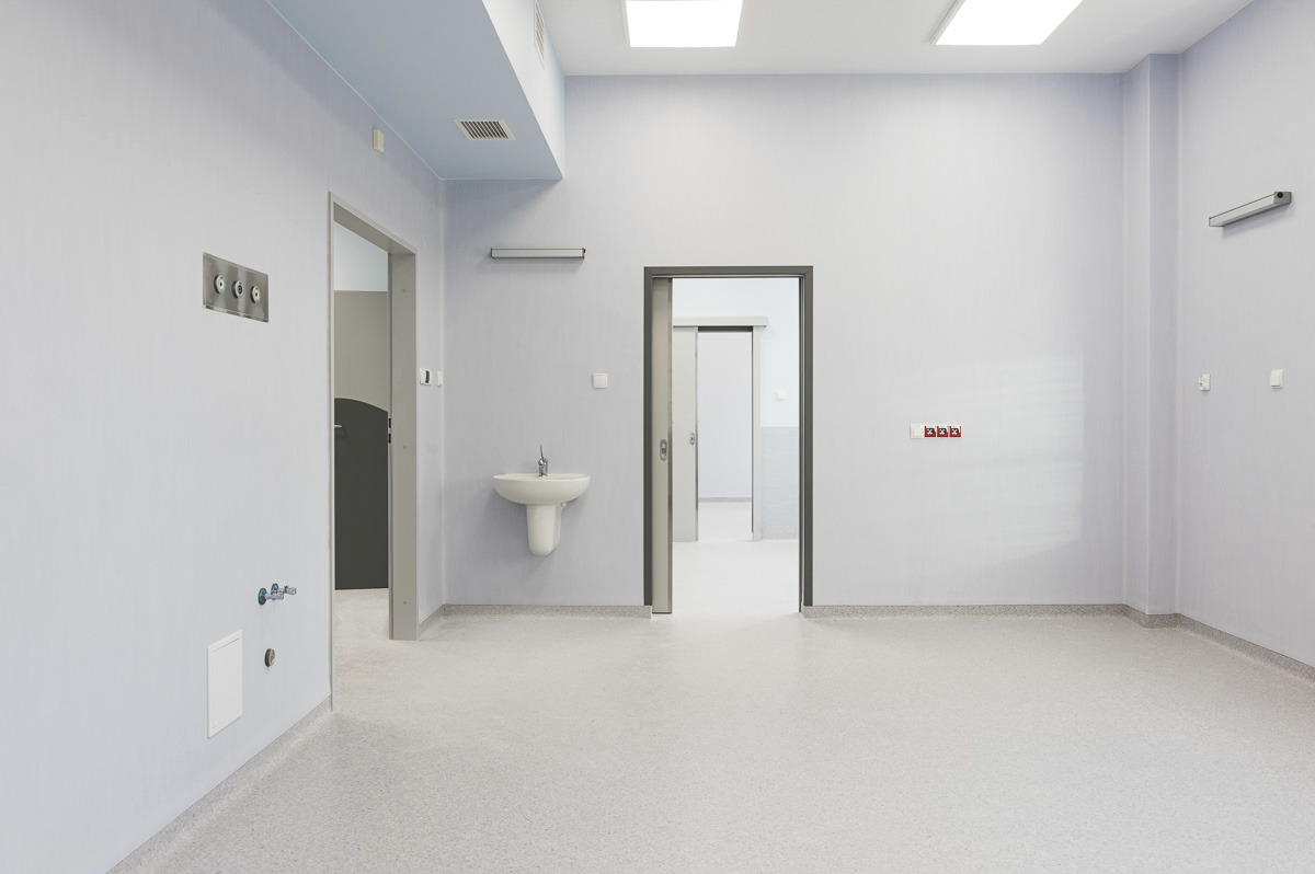 Puste, jasne pomieszczenie z szarą podłogą, umywalką na ścianie i kilkoma otwartymi drzwiami, oświetlone lampami sufitowymi.