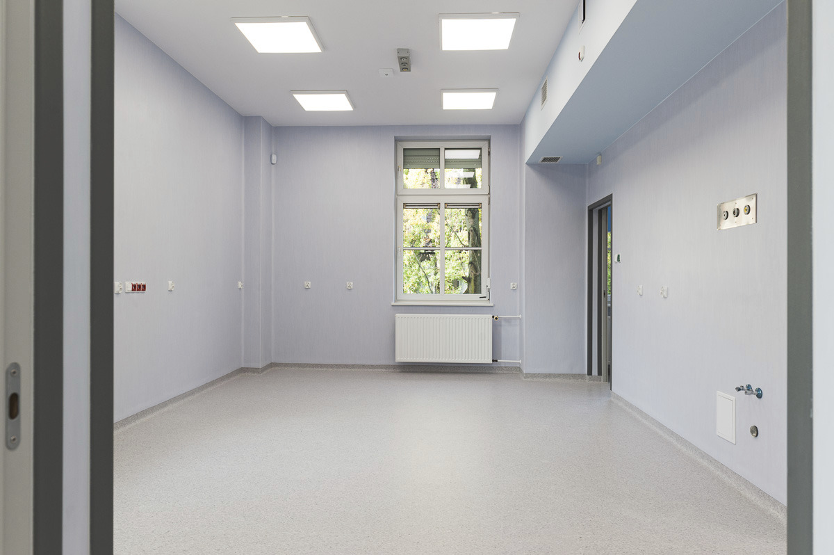 Puste, jasne pomieszczenie w odcieniach szarości z oknem, grzejnikiem i panelami ściennymi, oświetlone kwadratowymi lampami sufitowymi.