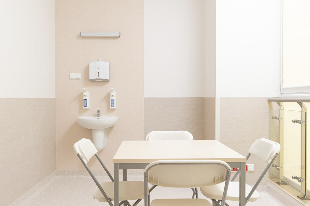 Jasne wnętrze z umywalką, dozownikami mydła, stołem i krzesłami; minimalistyczny design w odcieniach beżu i bieli.