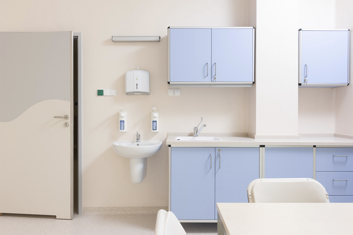 Jasne pomieszczenie z umywalką, szafkami w odcieniach błękitu i beżu, dozownikiem mydła na ścianie oraz częścią stołu i krzesła w dolnej części kadru, przypominające wnętrze gabinetu lekarskiego.
