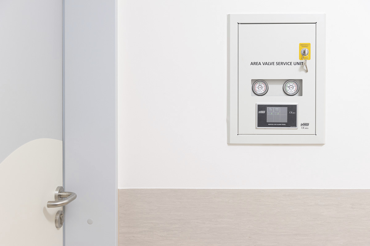 Biała skrzynka kontrolna z manometrami i panelem alarmowym gazów medycznych, zamontowana na ścianie w pomieszczeniu z szarą listwą przypodłogową i fragmentem drzwi z klamką po lewej stronie.
