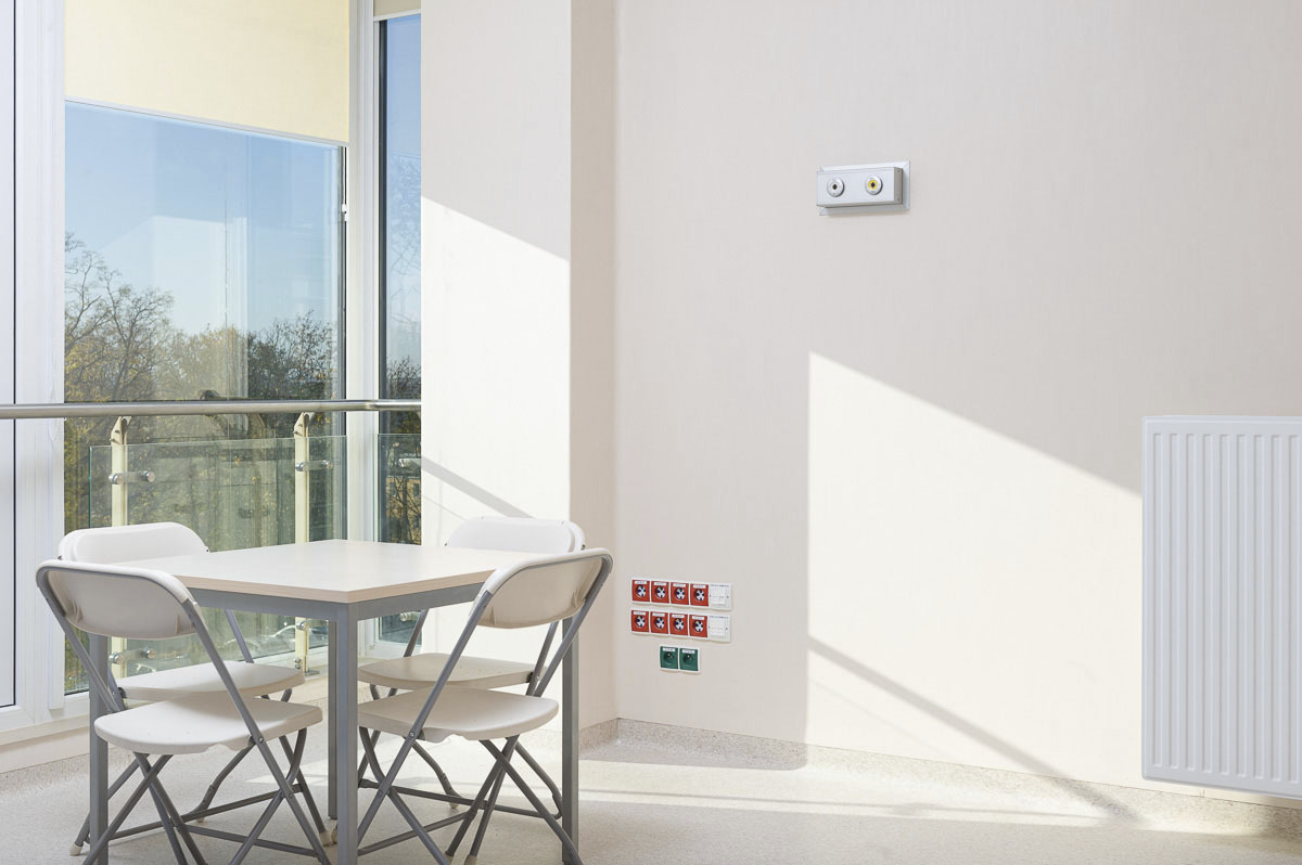 Jasne wnętrze z białym stołem i krzesłami, widok na balkon i drzewa za oknem, na ścianie gniazdka i przyciski alarmowe, grzejnik.