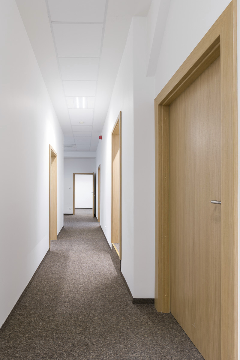 Długi korytarz biurowy z brązową wykładziną, białymi ścianami i jasnymi drewnianymi drzwiami. Widok w głąb korytarza z perspektywą zbieżną.