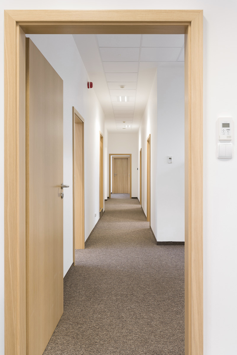 Wnętrze biurowego korytarza z brązową wykładziną, widok przez otwarte drewniane drzwi, białe ściany, oświetlenie sufitowe i drzwi po obu stronach korytarza.