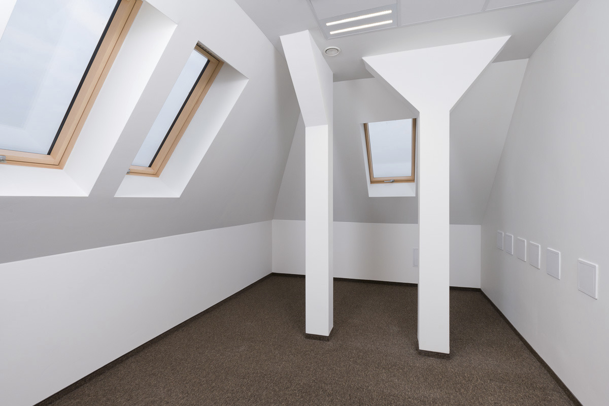 Puste, jasne poddasze z brązową wykładziną, trzema oknami dachowymi z drewnianymi ramami i białymi, nietypowymi filarami konstrukcyjnymi.
