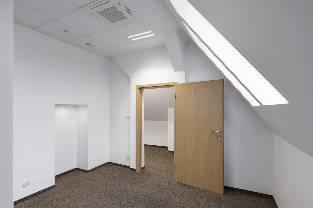 Puste, jasne pomieszczenie biurowe na poddaszu z oknem dachowym, brązową wykładziną i otwartymi drzwiami z jasnego drewna.