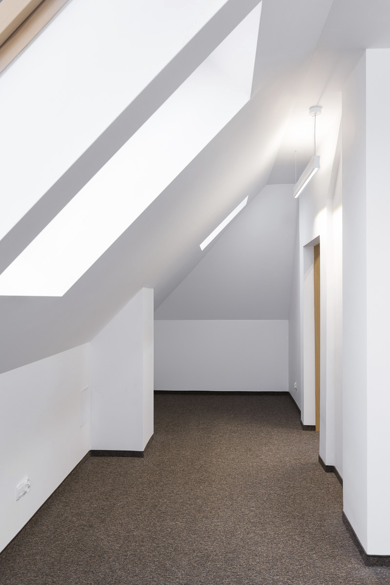Poddasze z oknami dachowymi, białymi ścianami i brązową wykładziną. W tle widoczna pusta przestrzeń. Oświetlenie stanowią podłużne lampy sufitowe.