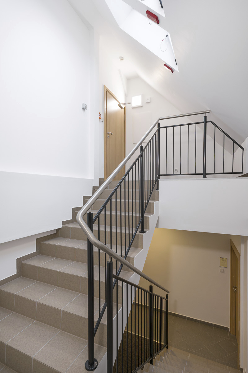 Klatka schodowa w budynku z beżowymi płytkami na schodach, metalowe czarne barierki i stalowa poręcz, białe ściany i drzwi na każdym piętrze.