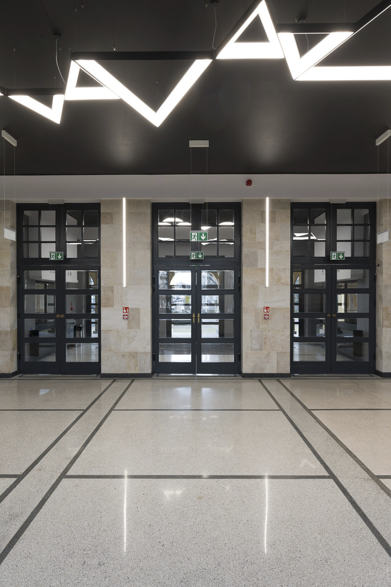 Wnętrze budynku z trzema parami przeszklonych drzwi, kamienną posadzką z geometrycznym wzorem i nowoczesnym oświetleniem sufitowym w kształcie zygzaków.