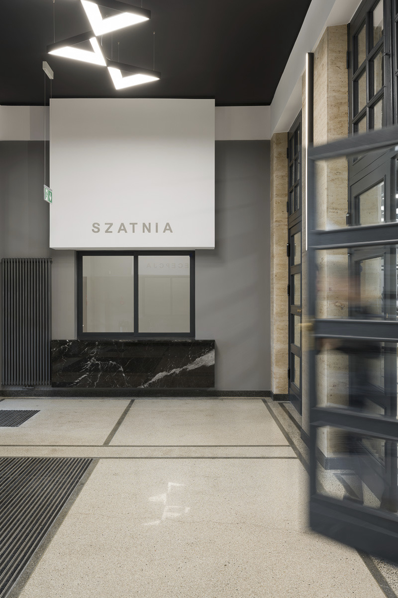 Wnętrze budynku z napisem 'SZATNIA' nad oknem recepcji, marmurowy blat, grzejnik żeberkowy, terakotowa podłoga z liniowymi wzorami, nowoczesne oświetlenie sufitowe, przeszklone drzwi z metalową ramą.