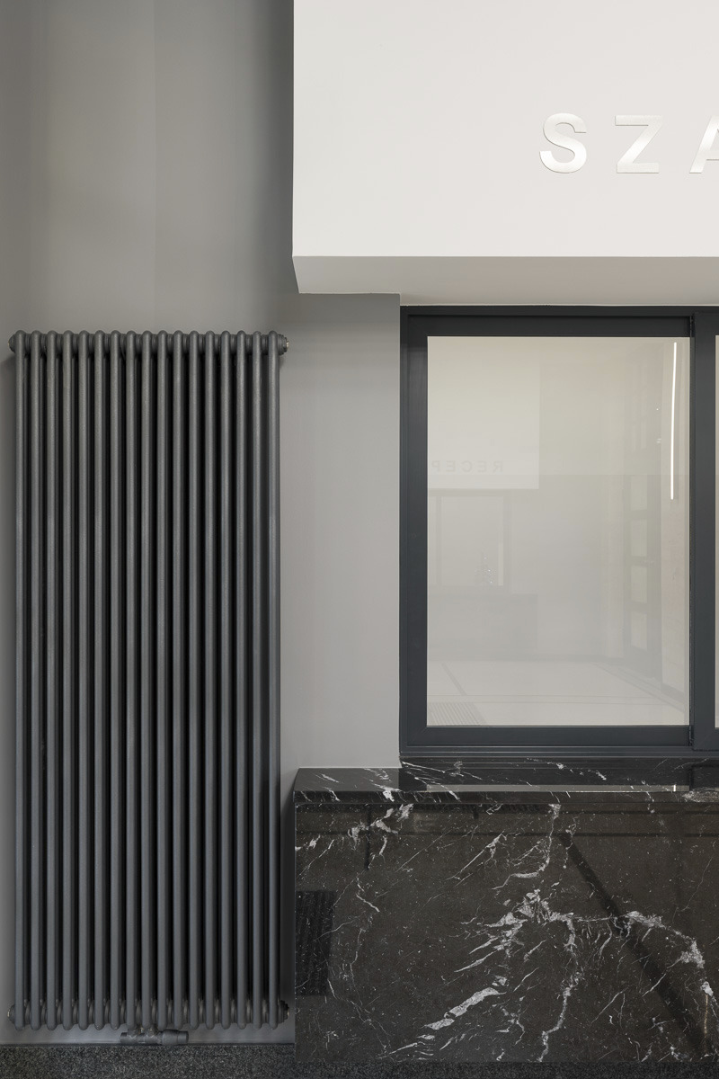 Szary grzejnik żeberkowy, czarny marmurowy parapet, okno z ciemną ramą i fragment napisu 'SZA' w jasnym wnętrzu.