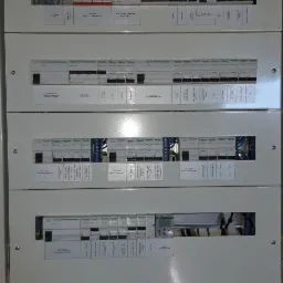 Rozdzielnica elektryczna z modułowymi wyłącznikami różnicowoprądowymi i nadprądowymi, opisana etykietami dla różnych obwodów, zamontowana na ścianie z surowej cegły.