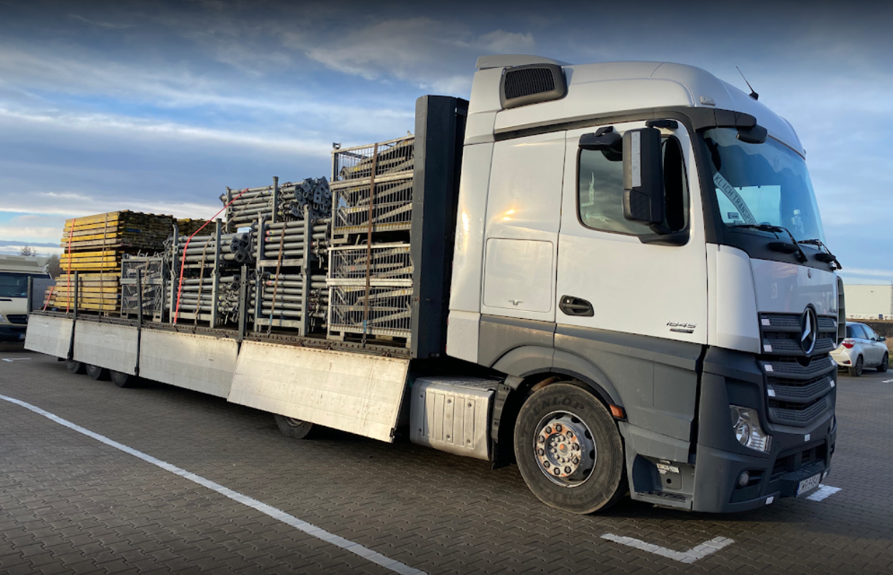 Ciężarówka Mercedes-Benz Actros załadowana metalowymi rusztowaniami i drewnianymi deskami, zabezpieczonymi czerwonymi pasami transportowymi, zaparkowana na parkingu.