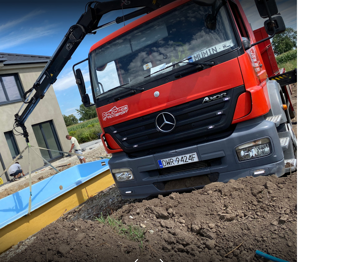 Czerwona ciężarówka z dźwigiem marki Mercedes-Benz Axor firmy transportowej Klichthos opuszcza niebieski basen do wykopu na tle nowego domu jednorodzinnego, widoczni dwaj pracownicy.