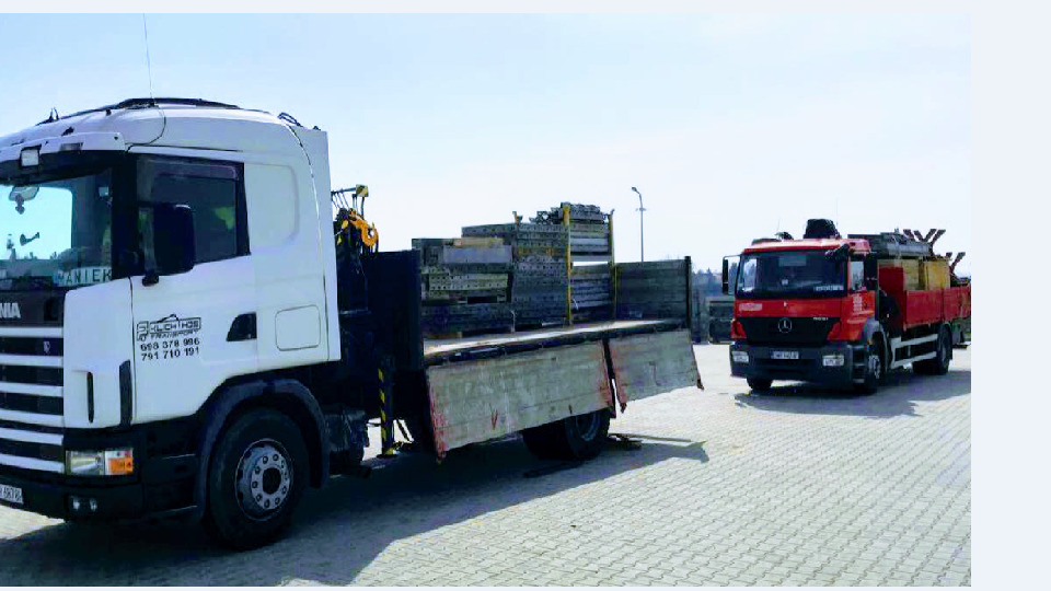 Dwa samochody ciężarowe: biały Scania z HDS i czerwony Mercedes, oba załadowane elementami szalunkowymi, stoją na szarym brukowanym placu w słoneczny dzień.